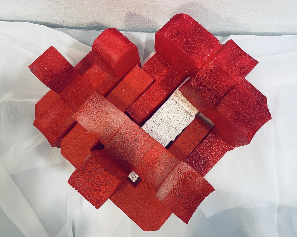 A red sculpture appearing from a foam like material. The sculpture is seen from above forming a heart like shape. It has different rectangular levels appearing in different block like shapes and heights. In the center is a small white area.