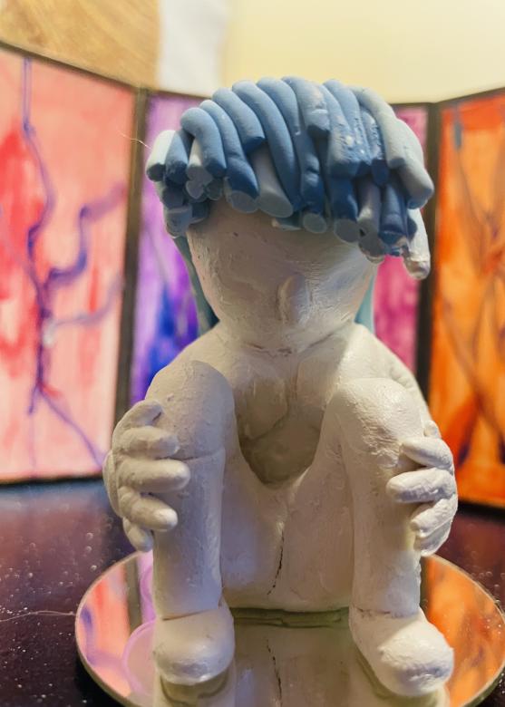 Close up of "The Internals" mixed media sculpture, the white clay figure is sitting on a circular mirrored surface holding its knees. The figure has minimal facial features and two toned blue hair hanging forward. Behind the figure is the colored glass.