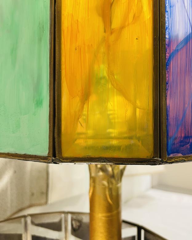 Close up of "The Internals" mixed media sculpture, through a yellow pane of glass you can make out the clay figure sitting inside. Below is the gold pole holding up the top layer of the sculpture. 