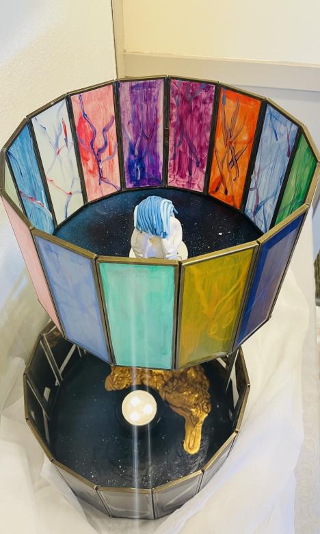 Mixed media sculpture with two circular layers made out of rectangular glass panes connected with a gold pole. The top layer, each pane of glass ic brightly colored but translucent; a small clay figure sits on the blue ground. On the bottom layer, inside of the clear glass panes is a light on the blue floor shining up;; gold paint spills around its side.