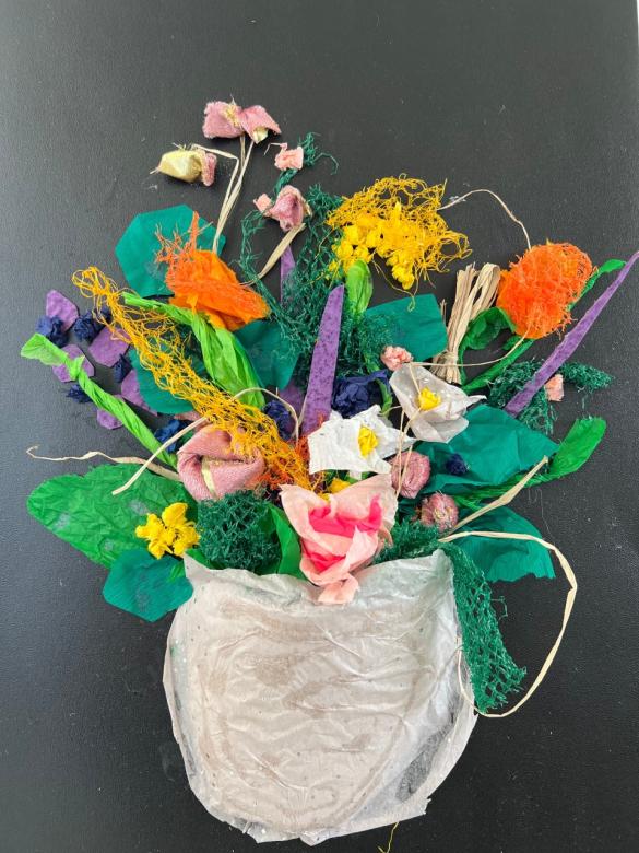 A textured paper/collaged bouquet of yellow, orange, pink, and purple flowers with green leaves in a white vase on a black background. The papers and fabrics making up the flowers include netting, tissue paper, and string.