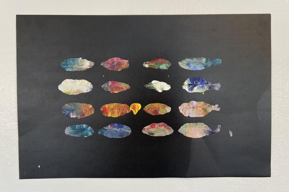 Black horizontal rectangular paper with acrylic paint printed on top. The print is aligned in the center forming a grid of colorful oval like shapes in a rectangle of four rows, four columns.