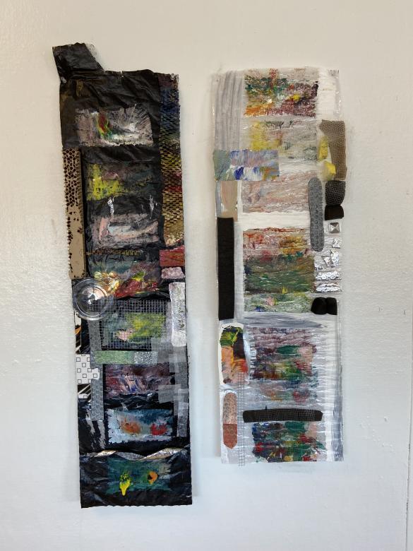 On a white wall, two thin rectangular shapes made from various recycled and found material hang parallel. On the right is a black rectangle, on the right is a white translucent rectangle. Colorful paint is present on each shape. 