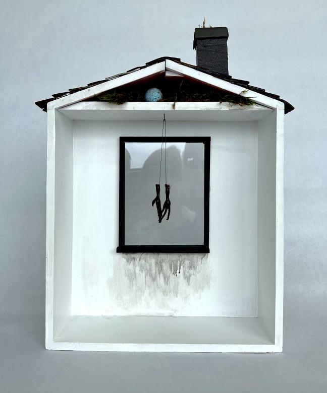 Sculpture of a rectangular white wooden house with the front open, a simple shingled roof, and black chimney. In the house is a black trimmed window with black paint smeared below. A black stick like object hangs in front of the window. In the attic appears moss and a blue robins egg.