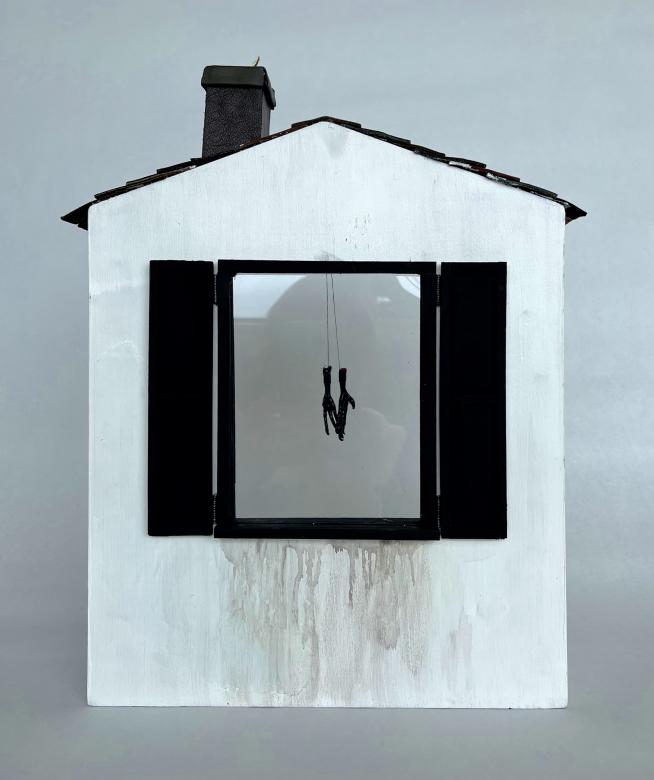 Sculpture of a white wooden house with a large window with black shutters. The house has black shingles and a black chimney. Black paint smears below the window. In the window appears two black stick like objects hanging.