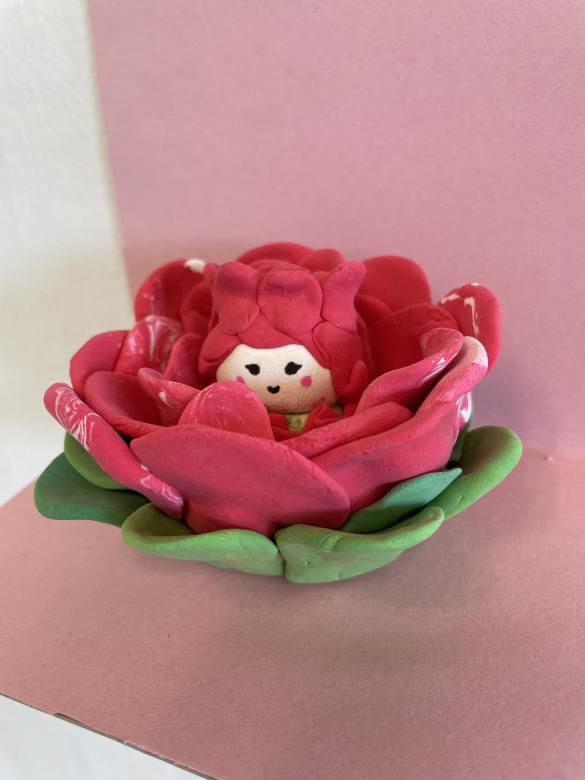 Model magic sculpture of a pink rose with green leaves. The rose is open in bloom with a small smiling rose creature. The sculpture is photographed on a light pink background. 