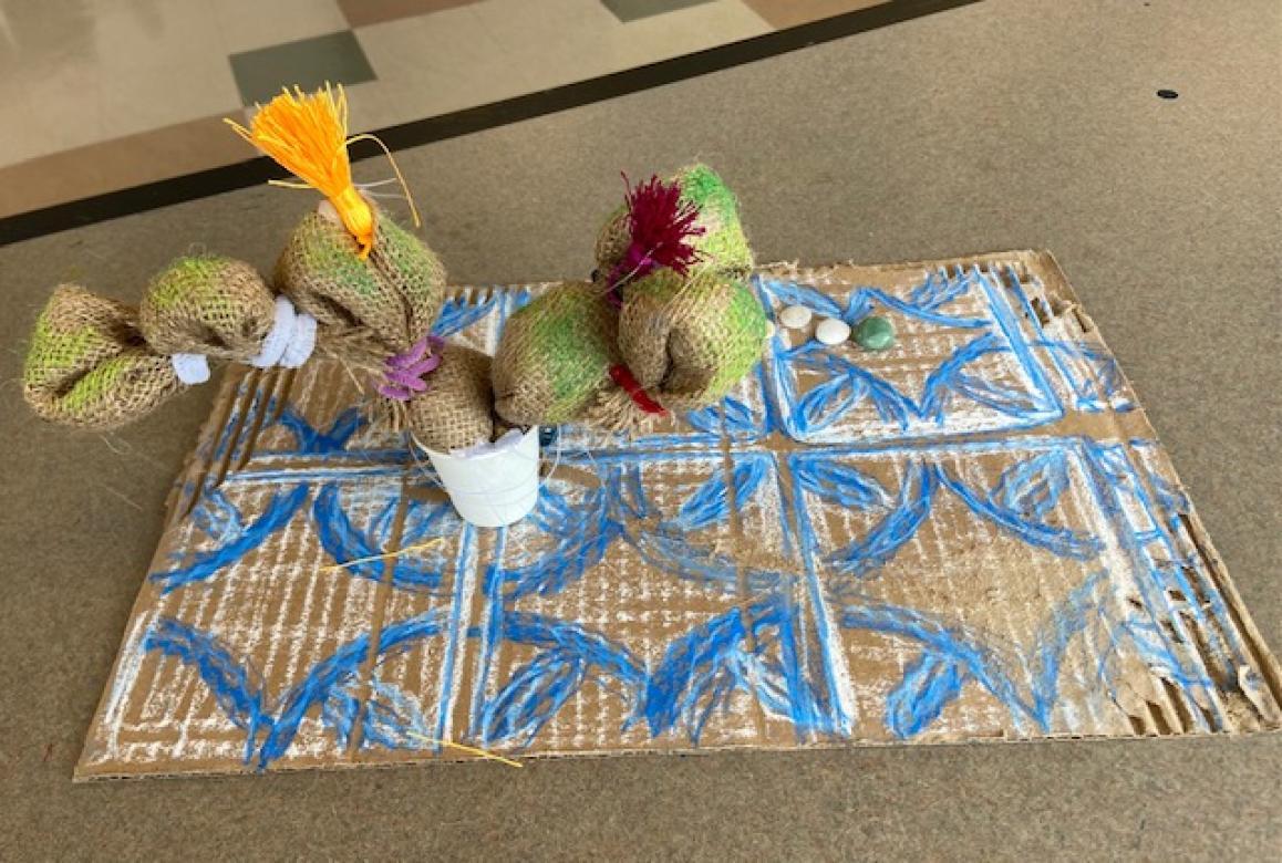 Sculpture of a cactus with magenta flowers made from fabric and recycled materials. It sits on piece of cardboard drawn with a blue and white vibrant tiled pattern.