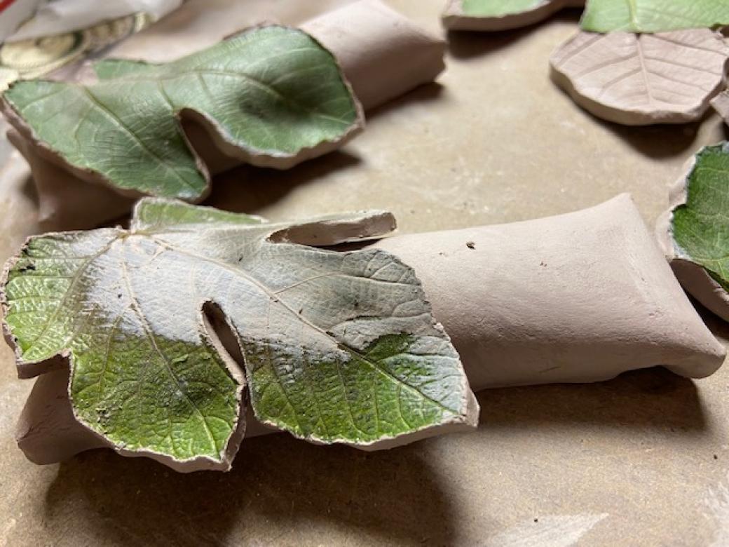 Photograph of a clay sculpture of fig leaves amongst rolls of clay. Some of the leaves are colored with shades of green.