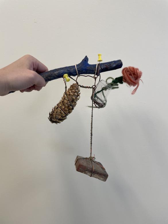 A hand holding a mobile sculpture. The hand is holding a blue branch which connects to hanging string containing small objects such as a pinecone, small box, and pipe cleaner flower.
