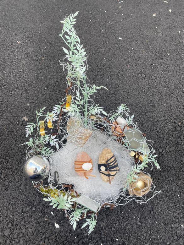 A mixed media sculpture of chicken wire and white tulle housing rocks and shells, The chicken wire extends upwards in the back wrapped with greenery, metallic objects, and brown pipe cleaners.  