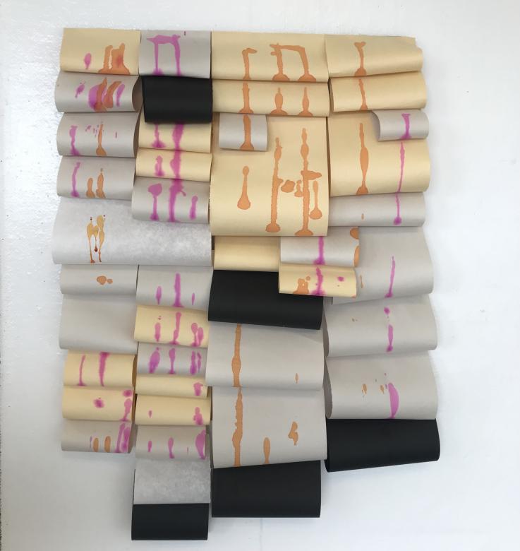 A wall hanging of rolls of off white, grey, yellow, and black small rolls of paper overlapping and cascading down. Pink and orange drips of ink adorn the paper.