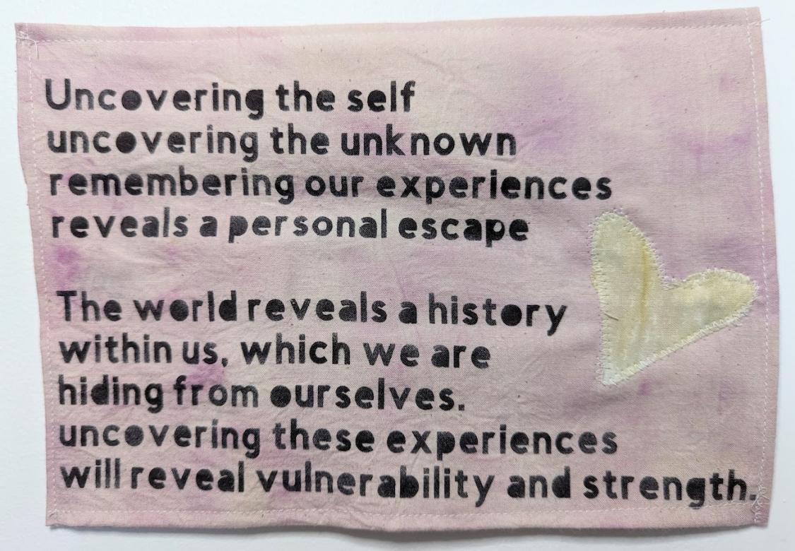 Pink piece of cloth with a yellow heart that reads in black text: Uncovering the self - uncovering the unknown - remembering our experiences - reveals a personal escape. The world reveals a history within us, which we are hiding from ourselves. Uncovering these experiences will reveal vulnerability and strength.