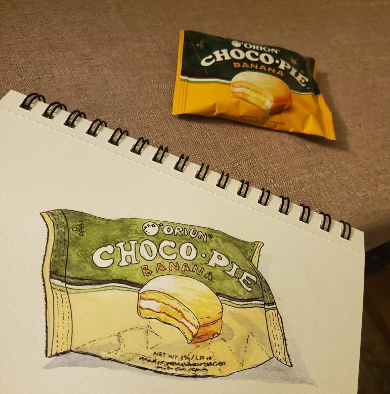 Photo of a choco pie in its wrapper next to a sketchbook with a drawing of the choco pie in wrapper