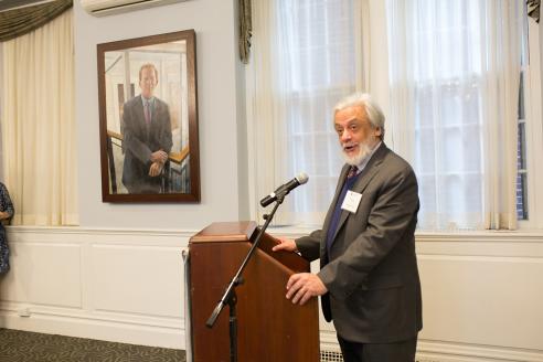 Tony Apesos speaks at a podium in Alumni Hall with his painting of Joe Moore visible in the background.