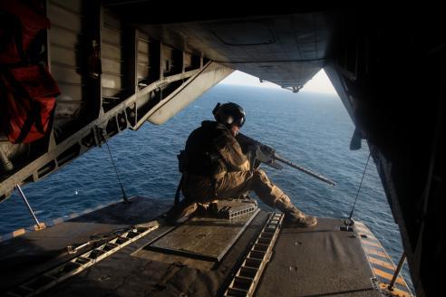 GULF OF OMAN, U.S. Marine Sgt. Gilbert Hopper provides security while flying aboard a CH-53E Super Stallion. Man crouched with gun on edge of open plane back with water in the background.