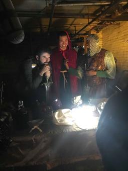 Three guys in a basement stand around a table wearing medevil knight costumes holding swords.