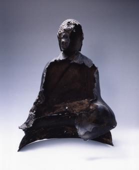 A photograph of a brown copper sculpture of a buddha sitting cross-legged that has been melted open and cracked apart in 