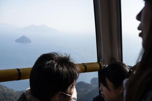 A photograph of mountains as seen from a cable car in Miyashima, Hiroshima. In the lower right corner, the obscured faces of one man and two women are seen looking out the cable car. 