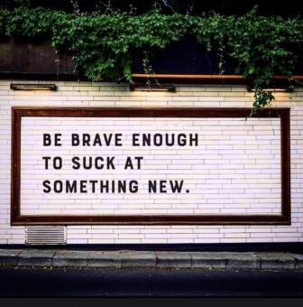 A sign on white brick that says "Be Brave Enough to Suck at Something New"