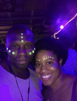 A photo of a black man (left) and black woman (right) in a dark room with purple neon lights. They both have face paint of green dots and blue stripes on their faces.