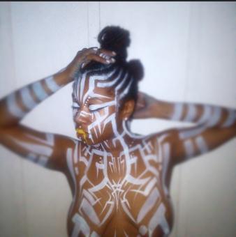 A topless black woman with white stripes of paint across her body.