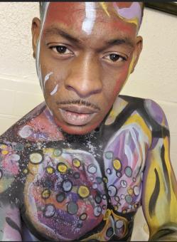 A photo of a black man with different colors of face paint on his body. Some of the colors include white, yellow, blue, purple, orange and grey.