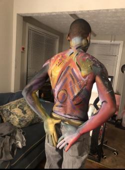 A photo of a black man, whose back is facing the photo, that has different colors of face paint on his body. Some of the colors include white, yellow, blue, purple, pink, orange and grey.