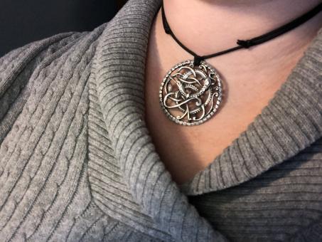 A silver circular pendant on a black cord, worn above the top half of a gray sweater.