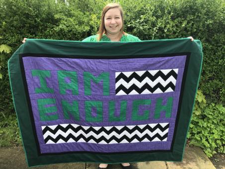 Person holding up a quilt with phrase “I am enough” in green with black/white chevron sections on a purple background with a black border, then dark green border, and bright green binding, in front of a green bush.