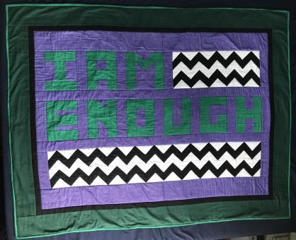 Quilt containing phrase "I am enough" in green including black/white chevron pattern on a purple background with a black border, then dark green border, then bright green binding. Dimensions 48 inches by 60 inches.