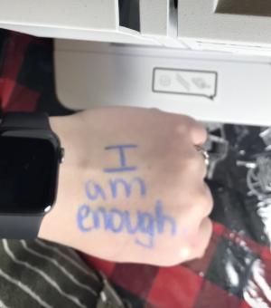 Hand with words "I am enough" written on it in blue body make up. Sewing machine in the top of the image with black/red plaid tablecloth underneath.