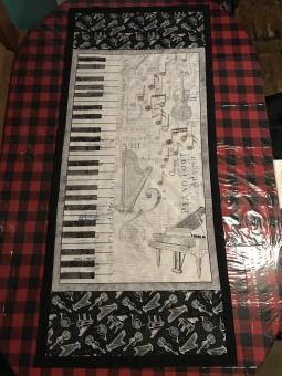 Music table runner including piano, harp, music note images with a black border on a red/black buffalo plaid table cloth. Dimensions 28 inches by 60 inches.