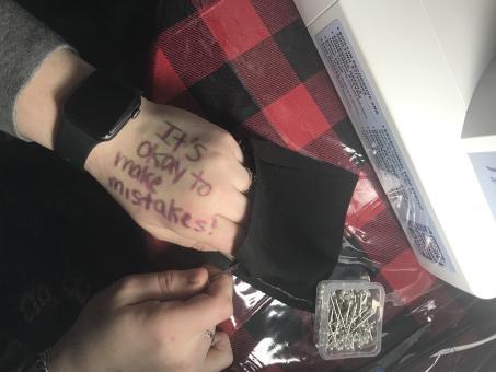 Two hands removing stitches from two black fabric pieces sewn together. One hand holds the tool. The other hand has "It's okay to make mistakes!" written on it in body make up in purple and green. Hands are shown on top of a black/red buffalo plaid tablecloth with a sewing machine on the table.