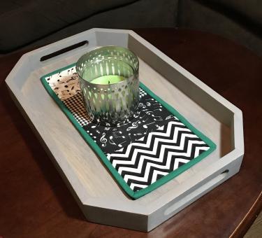 Quilted centerpiece including 4 blocks (white music note, buffalo plaid, black music note, and chevron fabric) binded with green fabric, sitting under a silver candle holder with a light green candle, on top of a grey tray on a coffee table.