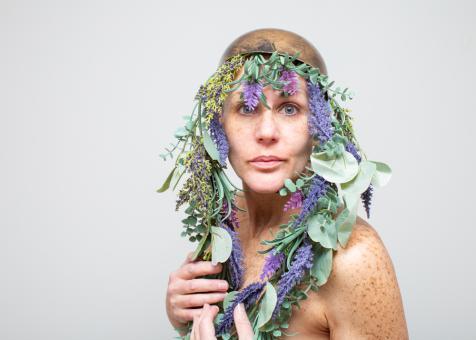 Photo of Eileen Powers wearing a brushed bronze cap with a garland of fake Lavender, Eucalyptus, Yellow and Purple Flowers, as well as other green across the front of the cap and hanging down each side of Eileen's face. Eileen clutches the garlands against her chest and looks at the viewer. 