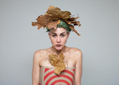 Photograph of Eileen Powers facing forward at the viewer from the waist up. She is wearing a headpiece made from large brown leaves and moss. She has on dark eye makeup and lipstick, she is clothed in a Target paper bag as a strapless dress. 