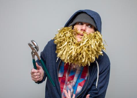 Photo of Eileen Powers holding a wrench in her left hand raised, hunched forward and looking to the right. Wearing a beanie, a navy sweatshirt with the hood pulled up, and a red, white, and blue flannel shirt. Eileen is wearing a voluminous beard and mustache made out of yellow gold tissue strands.