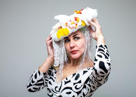 Photo of Eileen Powers facing to the left but looking to the right, holding a tilted headpiece on her head. The headpiece is an oval white shape, with white fabric and lace coming out on top and to the the left side. There are white daisies, yellow petals, and orange petals affixed to the top as well. Long irridescent white strands of easter grass plastic hang down on each side as hair. Eileen wears a patterned long sleeved black and white dress. 