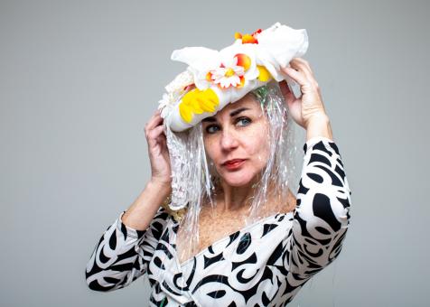 Photo of Eileen Powers facing to the left but looking to the right, holding a tilted headpiece on her head. The headpiece is an oval white shape, with white fabric and lace coming out on top and to the the left side. There are white daisies, yellow petals, and orange petals affixed to the top as well. Long irridescent white strands of easter grass plastic hang down on each side as hair. Eileen wears a patterned long sleeved black and white dress. 