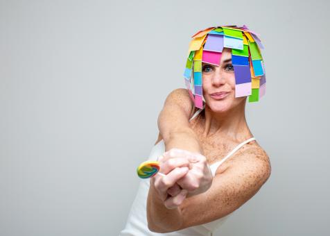 Photo of Eileen Powers wearing a white tank top, twisting and extending her arms towards the viewer holding a rainbow swirled lollipop. She is smiling and wearing a bob-length wig made of layered paint swatches in a rainbow of colors. 
