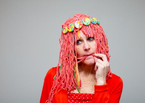 A photo of Eileen Powers dressed in a red long sleeve shirt with a red and white polka dot camisole. She is facing the camera with her head tilted, putting a sour rope strand into her mouth with her right hand. Her wig is made from red sour rope candies with a few strands of green, yellow, and orange sour rope candies. The wig has bangs with a headband made out of rainbow swirled lollipops.