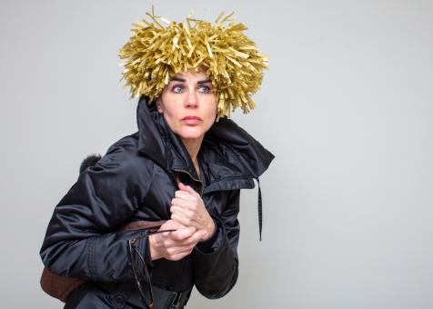 Image of Eileen Powers looking and facing to the right holding a bag and sunglasses with both hands. She is dressed in a navy blue jacket with a popped collar. She has a short wig sticking straight up made of golden yellow tissue strings.