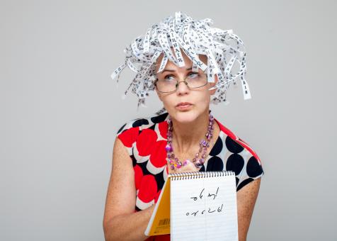 A photo of Eileen Powers looking up and to the left hunched forward. She is taking notes with a marker on a lined notepad. She has on a colorful black, white, and red circle print dress with a layered beaded necklace. She has on glasses with her lips pursed. Her hair is a short white wig made of ticker tape that repetitively spells hair over and over again in different fonts.   