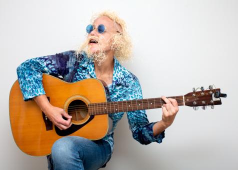 A Photo of Eileen Powers wearing a blue batik long sleeve button up, with jeans and round blue 60s style sunglasses. She is sitting facing the left holding and playing an acoustic guitar while singing.  She has medium length blonde wig made from straw packing material with an accompanying mustache and beard.
