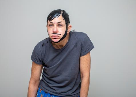 A photo of Eileen Powers wearing a grey short sleeve tshirt and lobster printed blue shorts.  She is hunched forward with her arms hanging down, she is squinting at the camera. She has short black stylized hair made from shiny black electrical tape. She also has a mustache, soul patch, and long sideburns that connect into a beard. 