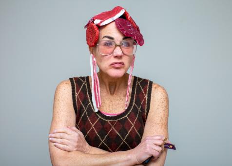 In this photo of Eileen Powers with arms crossed looking to the right scowling. She is wearing a black and neutral colored argyle sweater vest. She has on large vintage style glasses and is holding car keys in her hand. She has a short wig made of a a variety of plastic raw meats. She has a long plastic raw bacon earring in each ear. Her neutral pink lipstick matches the colors of the meats.