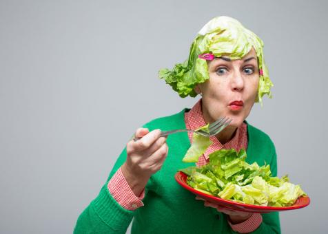 In this photo Eileen Powers in the right side of the image leaning forward looking left with her lips pursed chewing a piece of lettuce salad she is bringing towards her mouth with a fork. In the other hand she holds a red plate full of lettuce. She is wearing a red and white gingham collared shirt with a bright green cardigan over. She has her eyebrows raised and red lipstick on. Her wig is a bob made from large pieces of green lettuce with bangs and a hot pink hair clip pinned on each side.