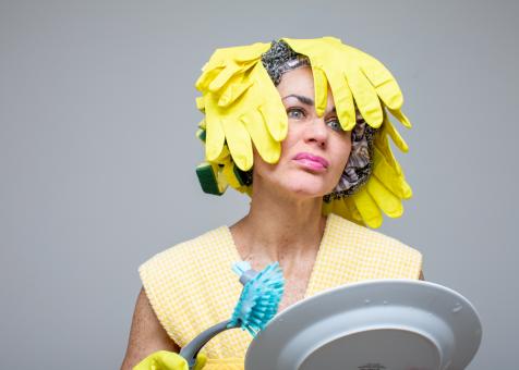 Photo of Eileen powers looking right aimed at the viewer, she is wearing a top made from yellow and white dish towels. She is also wearing yellow rubber dish gloves and scrubbing a plate with a blue soapy dish brush. Her wig is made from cascading yellow rubber dish gloves in some areas, with chunks of green and yellow sponges towards the back, and silver scrubbing pads along the front. 