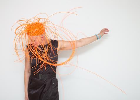 In this knee up portrait of Eileen Powers she is positioned to the left of the frame looking down holding her left arm up with an open hand. She is wearing shiny black cargo style pants with a matching belt and black tank top. She has on a multicolored thin beaded necklace, with three turquoise beaded bracelets on her raised hands. Her headpiece is a bright orange pillbox style cap fit snug with long orange plastic strings jutting out from every angle, some forming loops and swooping in front of her face.