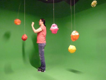 A student stands in the green-screened room with the hanging colored balls. The student's hand is outstretched as she had just pushed one of the balls.